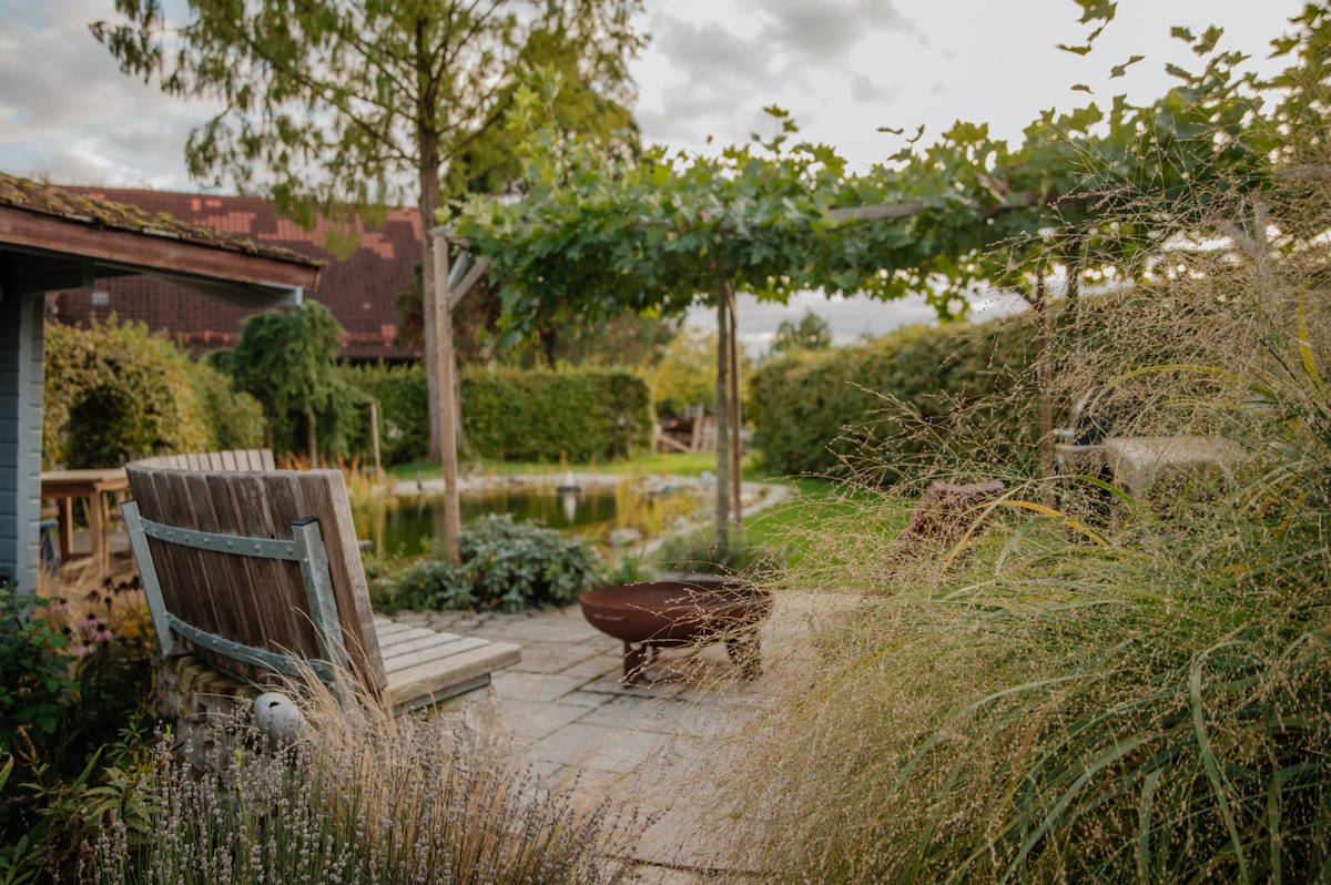 Gestalteter Sitzbereich im Garten mit Bepflanzung, im Hintergrund sieht man einen Naturteich