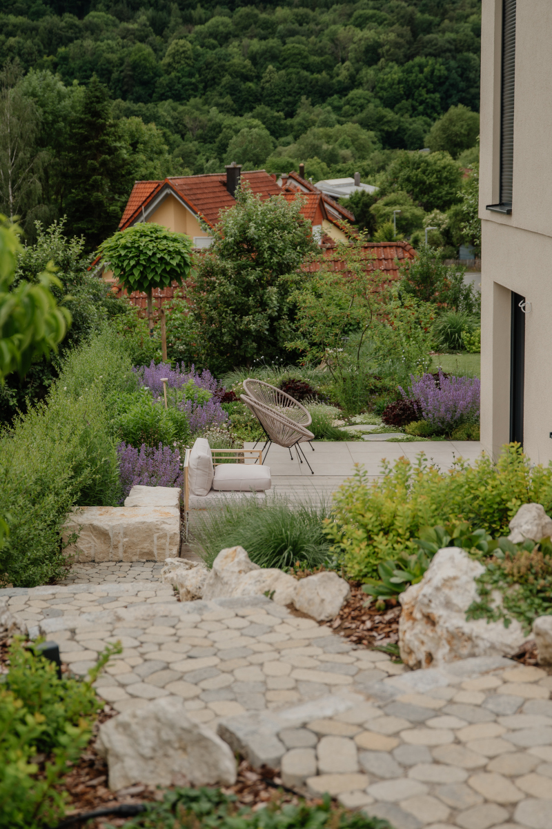 Blick von einer Natursteintreppe hinab auf eine moderne Terrasse mit Loungemöbeln im terrassierten Hanggarten.